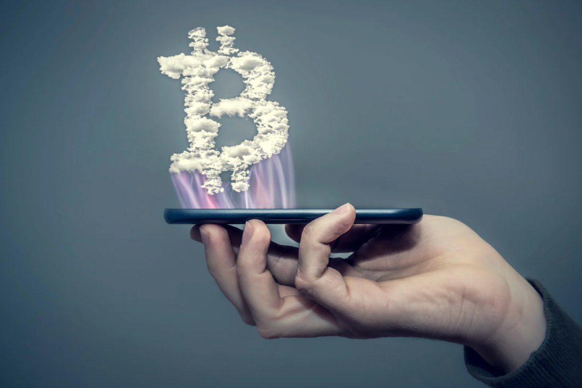 mano che regge uno smartphone da cui esce la B bianca di Bitcoin