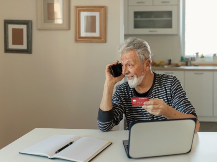 Anziano al telefono con il bancomat in mano, davanti al computer